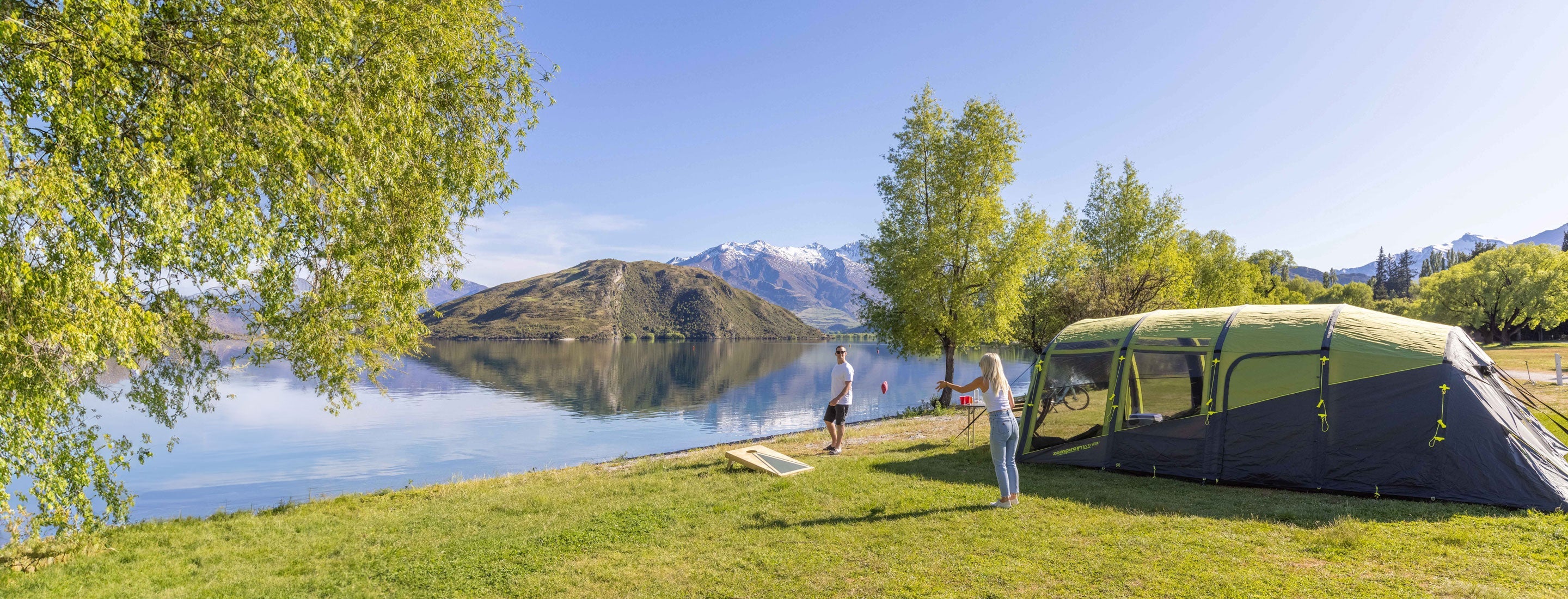 Two people near a tent by a lake with mountains in the background