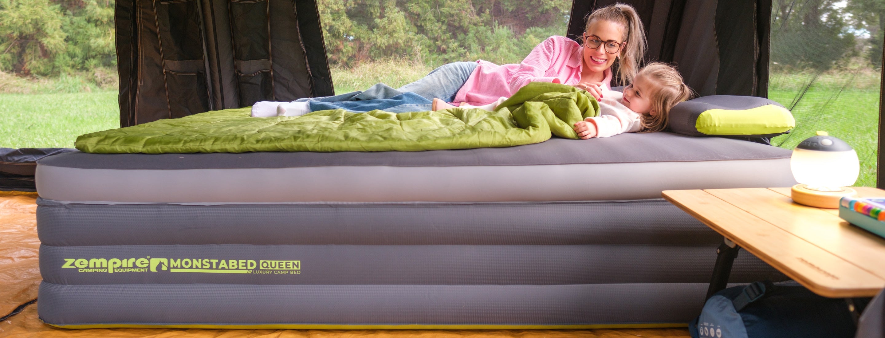 Woman and child lying on a Zempire Monstabed Queen air mattress in a camping setting.