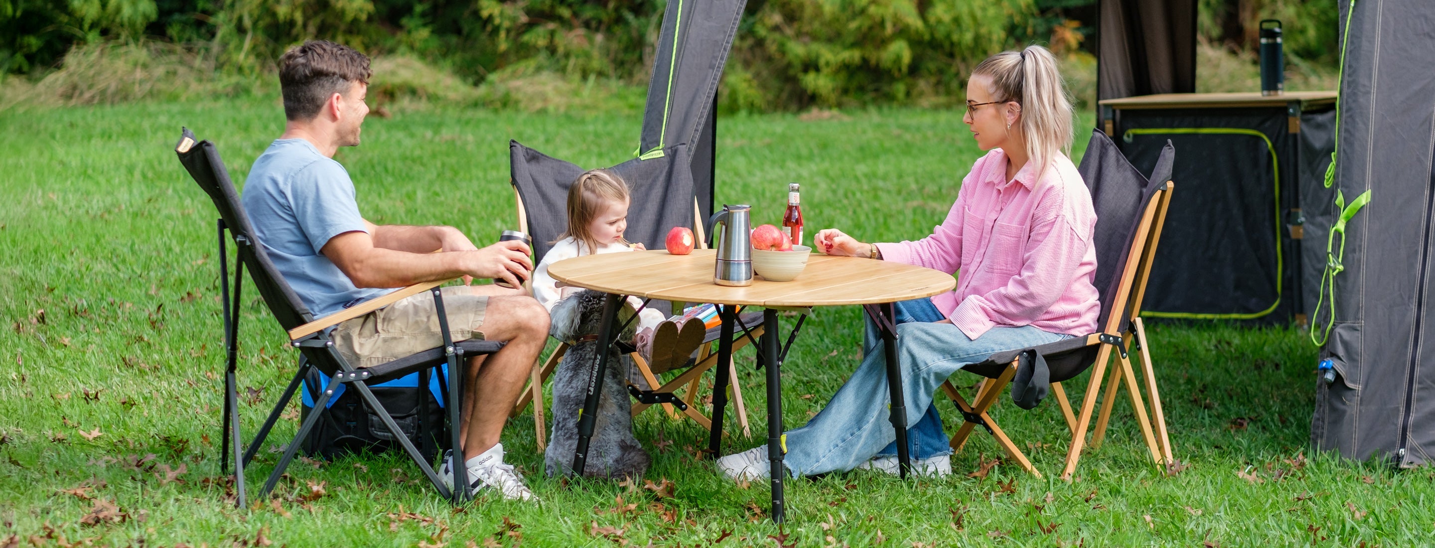 Family of three sitting at a table outdoors with camping gear around
